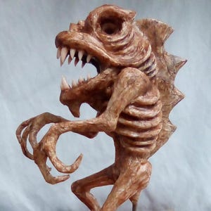 May include: A detailed sculpture of a creature with a reptilian head, sharp teeth, and a skeletal torso. The sculpture is primarily brown with textured details, featuring clawed hands and a spiky back. The creature appears to be in a dynamic pose.