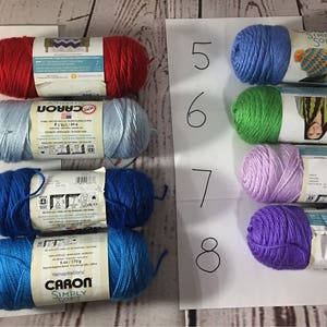 Puede incluir: Ocho ovillos de lana en varios colores, incluyendo rojo, azul claro, azul oscuro y verde. La lana est&aacute; etiquetada con la marca "Caron Simply Soft" y est&aacute; dispuesta sobre una superficie blanca. Cada ovillo pesa aproximadamente 170 gramos.