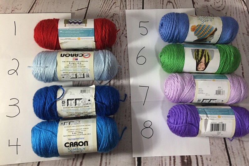 Puede incluir: Ocho ovillos de lana en varios colores, incluyendo rojo, azul claro, azul oscuro y verde. La lana est&aacute; etiquetada con la marca "Caron Simply Soft" y est&aacute; dispuesta sobre una superficie blanca. Cada ovillo pesa aproximadamente 170 gramos.