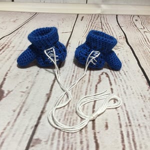 Puede incluir: Un par de botines de beb&eacute; azules de ganchillo hechos a mano con cordones blancos. Los botines se muestran sobre una superficie de madera de color claro. Los cordones est&aacute;n atados.