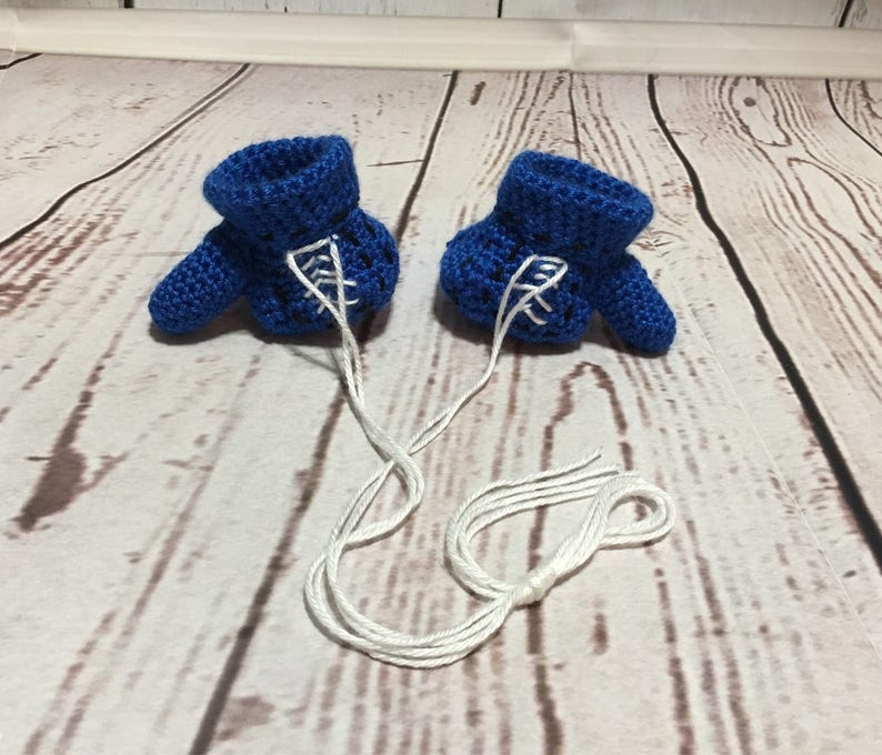 Puede incluir: Un par de botines de beb&eacute; azules de ganchillo hechos a mano con cordones blancos. Los botines se muestran sobre una superficie de madera de color claro. Los cordones est&aacute;n atados.