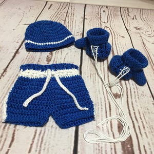 Puede incluir: Un conjunto de boxeo para beb&eacute; tejido a crochet en azul real con detalles blancos. El conjunto incluye un gorro, pantalones cortos con cord&oacute;n y guantes de boxeo. El gorro tiene una raya blanca. Los pantalones cortos tienen ribetes blancos. Los guantes tienen cordones blancos.