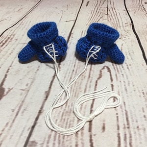 Puede incluir: Un par de patucos de beb&eacute; azules de ganchillo hechos a mano con detalles blancos y cordones de hilo blanco adjuntos. Los patucos se muestran sobre una superficie de vetas de madera clara.