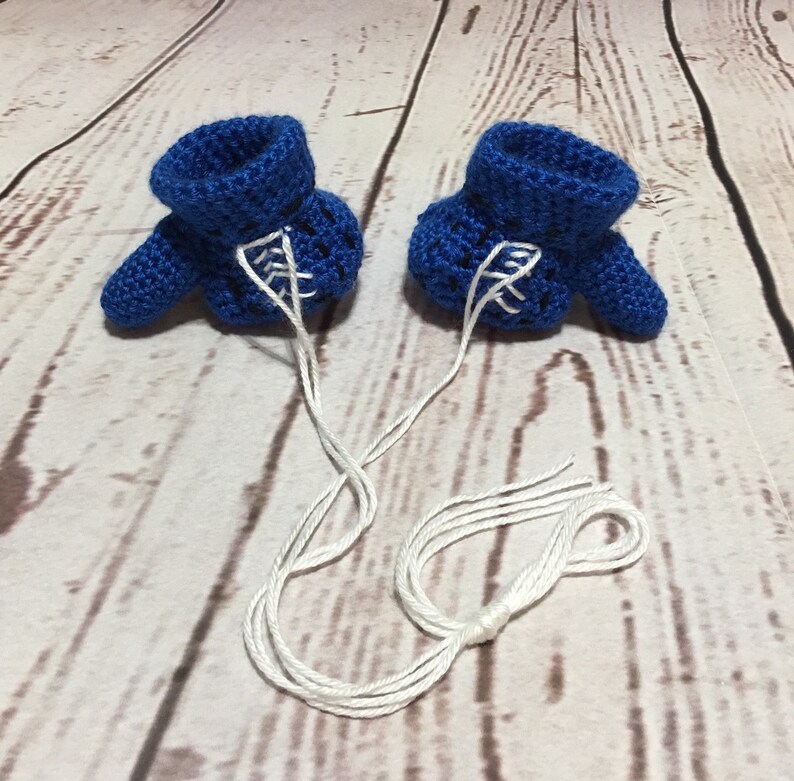 Puede incluir: Un par de patucos de beb&eacute; azules de ganchillo hechos a mano con detalles blancos y cordones de hilo blanco adjuntos. Los patucos se muestran sobre una superficie de vetas de madera clara.