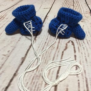 Puede incluir: Dos patucos de beb&eacute; azules de ganchillo hechos a mano con cordones blancos. Los patucos est&aacute;n conectados por una cuerda blanca y se muestran sobre una superficie de vetas de madera clara. Los patucos tienen un pu&ntilde;o y una peque&ntilde;a puntera.