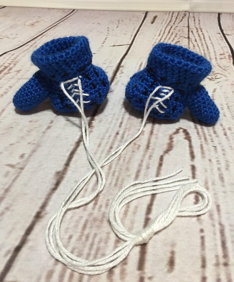 Puede incluir: Dos patucos de beb&eacute; azules de ganchillo hechos a mano con cordones blancos. Los patucos est&aacute;n conectados por una cuerda blanca y se muestran sobre una superficie de vetas de madera clara. Los patucos tienen un pu&ntilde;o y una peque&ntilde;a puntera.