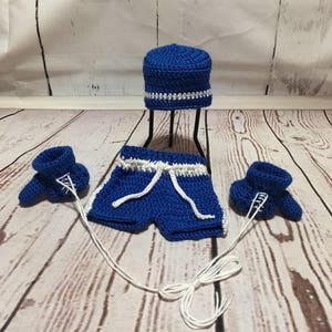 Puede incluir: Un conjunto de ganchillo hecho a mano en azul real con detalles en blanco, que incluye pantalones cortos con cord&oacute;n, un gorro y guantes de boxeo. Los guantes est&aacute;n conectados a los pantalones cortos con hilo blanco. El gorro est&aacute; sobre un soporte negro.