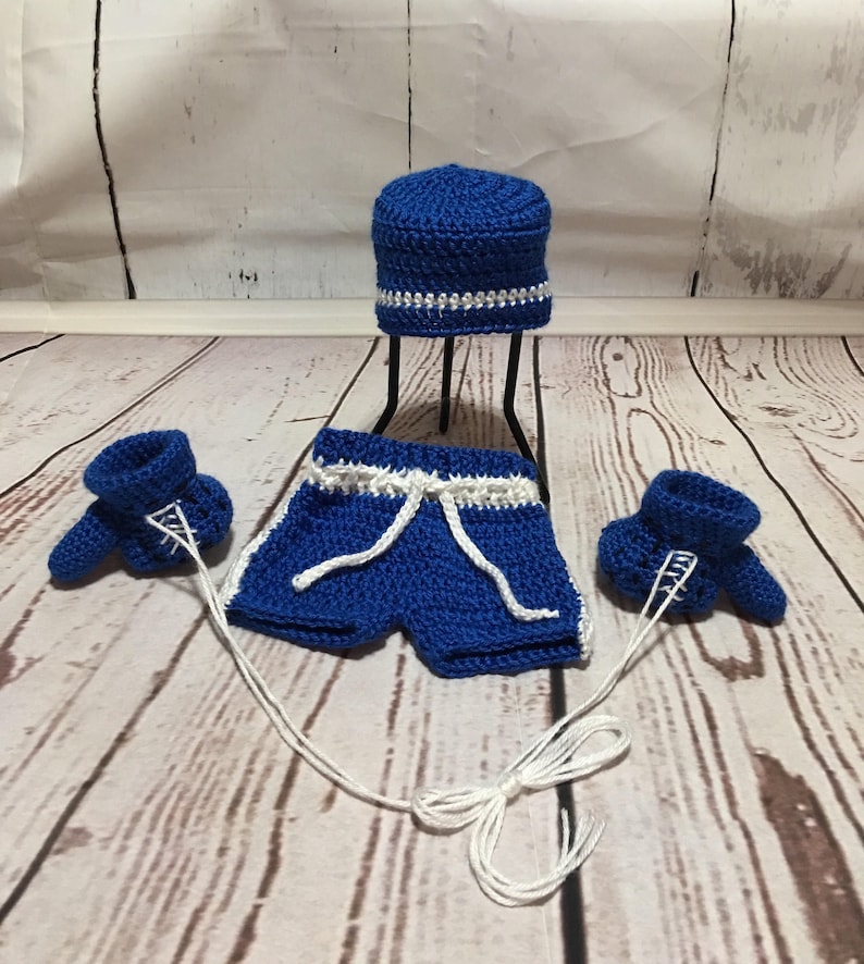 Puede incluir: Un conjunto de ganchillo hecho a mano en azul real con detalles en blanco, que incluye pantalones cortos con cord&oacute;n, un gorro y guantes de boxeo. Los guantes est&aacute;n conectados a los pantalones cortos con hilo blanco. El gorro est&aacute; sobre un soporte negro.