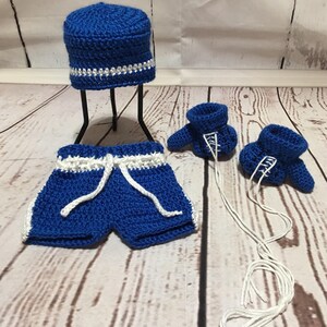 Puede incluir: Un conjunto de beb&eacute; de ganchillo hecho a mano en azul real con detalles blancos. El conjunto incluye un gorro, pantalones cortos con cord&oacute;n y dos patucos. El gorro tiene un ribete blanco y plateado. Los pantalones cortos y los patucos tienen ribete blanco.