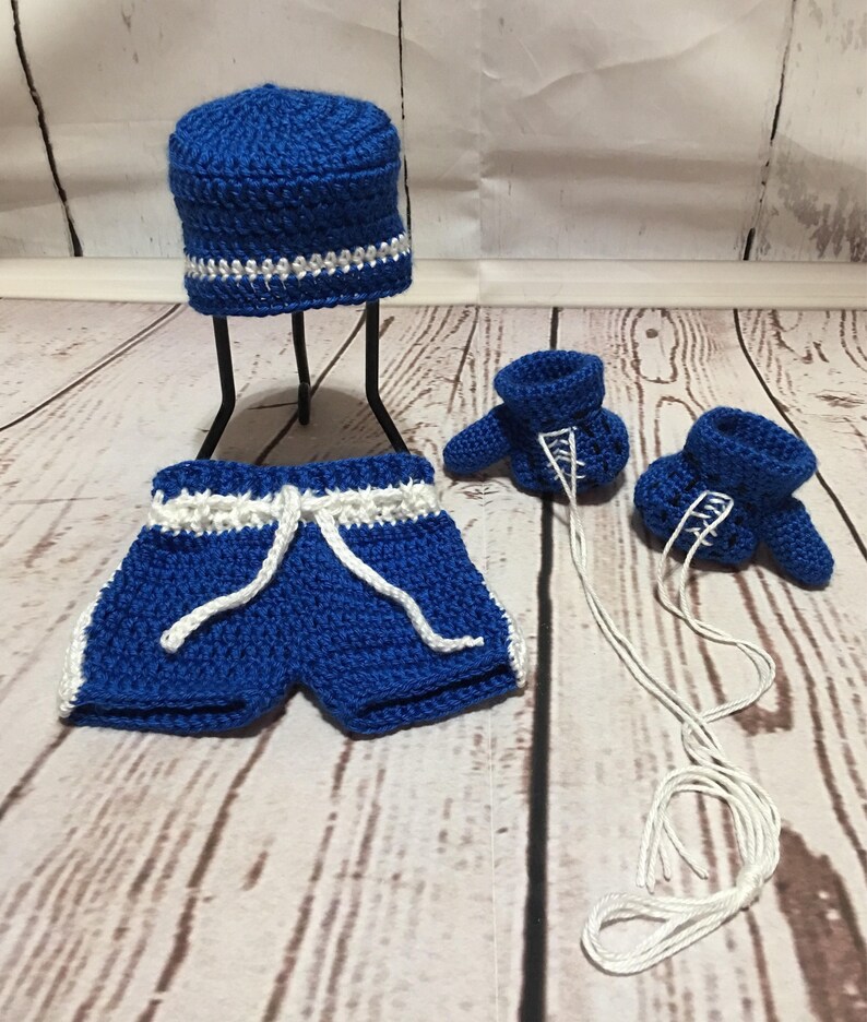 Puede incluir: Un conjunto de beb&eacute; de ganchillo hecho a mano en azul real con detalles blancos. El conjunto incluye un gorro, pantalones cortos con cord&oacute;n y dos patucos. El gorro tiene un ribete blanco y plateado. Los pantalones cortos y los patucos tienen ribete blanco.