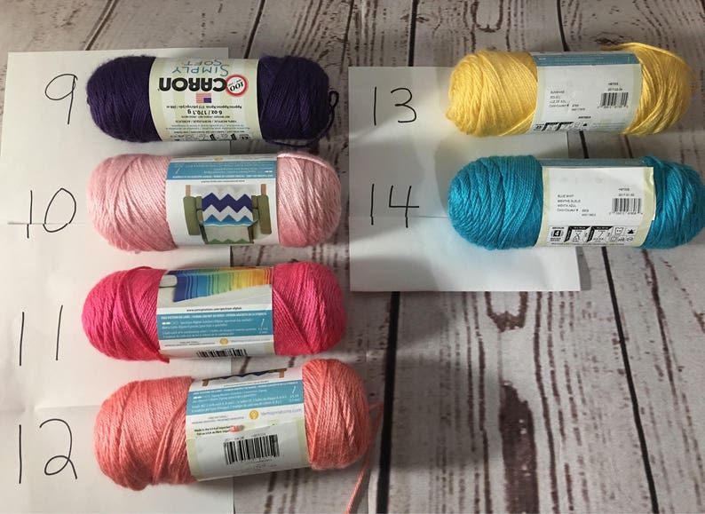 Puede incluir: Surtido de ovillos de hilo Caron Simply Soft en varios colores, incluyendo morado, rosa, coral, amarillo y turquesa. El hilo est&aacute; dispuesto sobre una superficie clara con vetas de madera, con etiquetas de papel blanco numeradas.