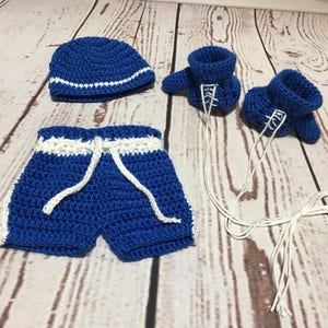 Puede incluir: Conjunto de beb&eacute; de ganchillo azul con detalles blancos. El conjunto incluye un gorro, un pantal&oacute;n corto con cord&oacute;n y patucos con cordones blancos. Presentado sobre una superficie de imitaci&oacute;n de madera clara.