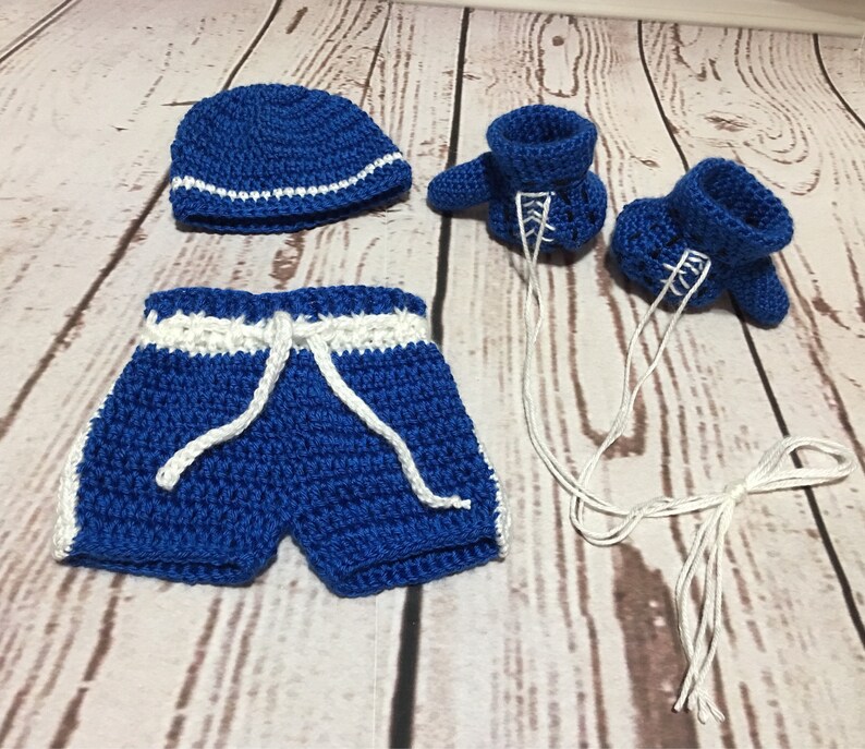 Puede incluir: Conjunto de beb&eacute; de ganchillo azul con detalles blancos. El conjunto incluye un gorro, un pantal&oacute;n corto con cord&oacute;n y patucos con cordones blancos. Presentado sobre una superficie de imitaci&oacute;n de madera clara.