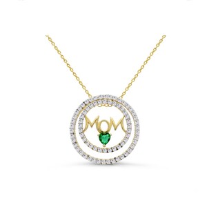 Può includere: Collana d'oro con un ciondolo circolare che presenta la parola "MOM" in lettere corsive con una pietra preziosa verde a forma di cuore al centro. Il ciondolo è circondato da due file di pietre scintillanti trasparenti.