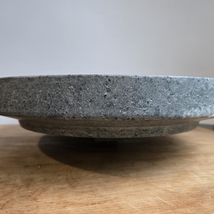 May include: A gray, stone bowl with a wide, flat rim and a slightly raised base. The bowl has a rough, textured surface and sits on a wooden surface. The bowl's design is simple and functional.