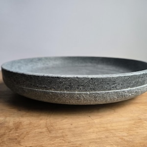 May include: A gray stone bowl with a wide, shallow design. The bowl has a textured surface and a lighter-colored band around the base. It sits on a wooden surface.