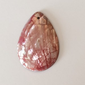 May include: A pink and white pearlescent shell pendant with a hole at the top for stringing. The shell is cracked and has a mosaic-like appearance.