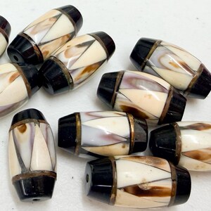 May include: A collection of barrel-shaped beads. Each bead features a cream-colored central section with brown and purple accents, framed by black end caps and a metallic band. The beads are scattered across a white surface.