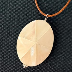 May include: An oval-shaped pendant crafted from iridescent mother-of-pearl, featuring a segmented starburst pattern on its surface. The pendant is suspended from a brown leather cord and includes a small silver-toned metal accent at the base.