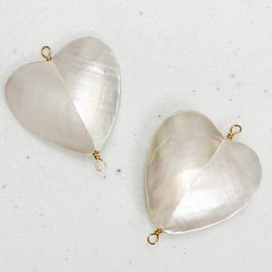 May include: Two heart-shaped pendants with a pearlescent sheen. Each pendant is divided diagonally, with a gold-colored wire loop at the top and bottom for hanging. The pendants are set against a light background.