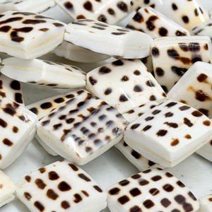 May include: A collection of square, white beads with irregular brown spots. The beads have rounded edges and a glossy finish, resembling a natural stone or shell. These beads are suitable for jewelry making.