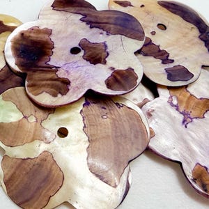 May include: A collection of six flower-shaped wooden buttons. The buttons have a light beige base color with brown and purple-hued patterns. Each button has a small hole in the center for sewing. The buttons are arranged in a slightly overlapping pattern.