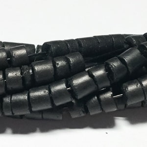 May include: Black wooden beads with a cylindrical shape and a grooved texture. The beads are stacked together in a pile.