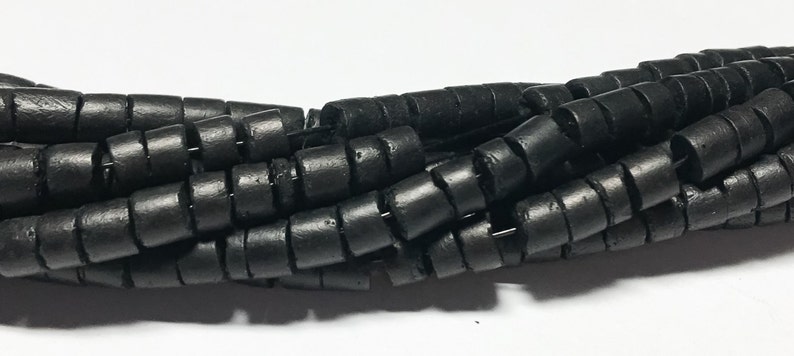 May include: Black wooden beads with a cylindrical shape and a grooved texture. The beads are stacked together in a pile.