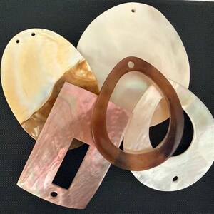May include: A collection of assorted jewellery components made from iridescent shell material. The set includes oval, rectangular, and teardrop-shaped pieces in shades of cream, brown, and soft pink, each featuring small pre-drilled holes for attachment.