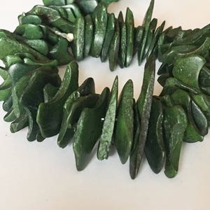 May include: A dark green bracelet composed of carved, irregular pieces. The bracelet's components are strung together, forming a circular shape. The pieces have a polished appearance, and the overall design is organic and natural.