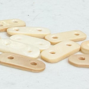 May include: A collection of small, rectangular, off-white wooden beads with two holes each. The beads vary slightly in color, ranging from a pale cream to a light tan. They are scattered on a white surface.