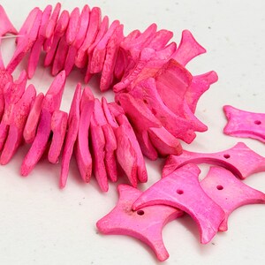 以下が含まれることがあります： 明るいピンク色の、彫刻された木製ビーズで作られたネックレス。ビーズは不規則な形をしており、一緒に紐で繋がれています。いくつかのマッチするピンク色の星型のビーズが近くに散らばっています。ネックレスとビーズは白い表面にあります。