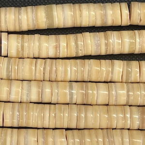 Puede incluir: Primer plano de varios hilos de cuentas beige planas en forma de disco. Las cuentas tienen un aspecto natural y orgánico con sutiles variaciones de color y textura. Las cuentas están ensartadas, creando una superficie texturizada. El fondo es de color oscuro.