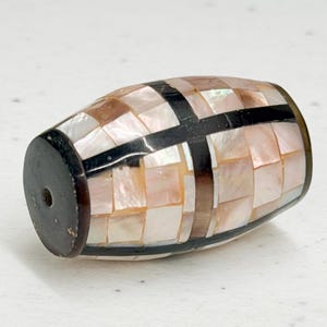 May include: A decorative barrel-shaped bead. The bead is made of small, square, iridescent mother-of-pearl tiles in shades of pink, cream, and brown. Black bands and end caps add contrast. A small hole is visible on one end.