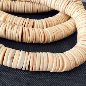 Könnte beinhalten: Ein langer Strang aus cremefarbenen, kreisförmigen, flachen scheibenförmigen Perlen, die in einer durchgehenden Spirale angeordnet sind. Jede einzelne Perle hat eine glatte, matte Oberfläche mit subtilen Farbnuancen für ein einheitliches Bild.