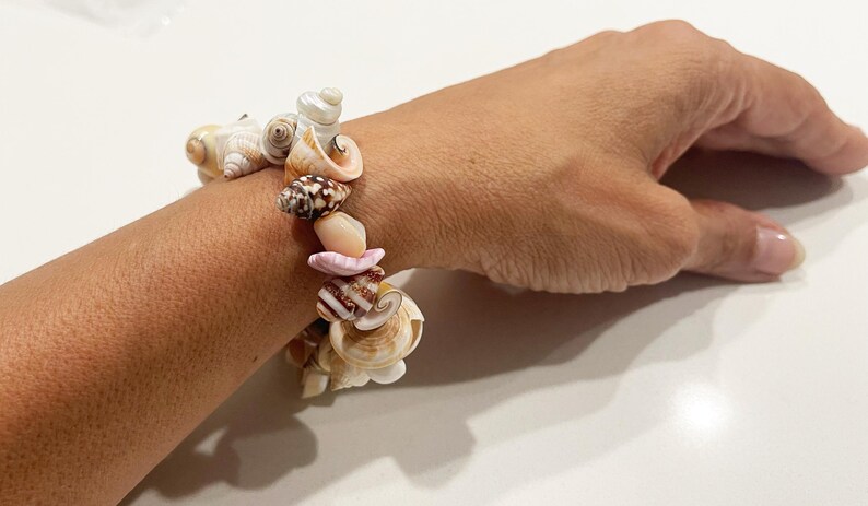 May include: A bracelet composed of various natural seashells in shades of white, tan, brown, and soft pink. The shells are arranged in a cluster, featuring diverse spiral and conical shapes, secured together to form a circular wrist accessory.