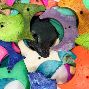 May include: A collection of crescent moon-shaped pendants in various colors including lime green, black, purple, orange, blue, pink, and white. Each piece features a textured surface and a small circular hole for threading, arranged in a dense pile.