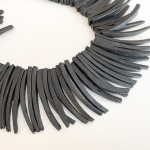 May include: A black necklace composed of numerous curved, rectangular pieces. The pieces are arranged in a circular pattern, creating a fringe-like effect. The necklace is set against a white background, highlighting its dark color and unique design.