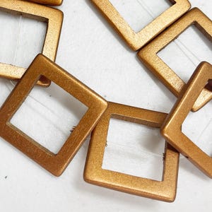 May include: Close-up of a necklace featuring square, open-frame beads in a metallic bronze color. The beads are strung together with a thin, clear cord, creating a geometric pattern. The necklace is displayed on a white surface.