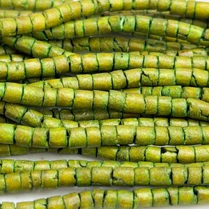 May include: Close-up of several strands of green beads. The beads are cylindrical with a square cross-section and have a textured, weathered appearance. The color is a mix of olive and chartreuse.