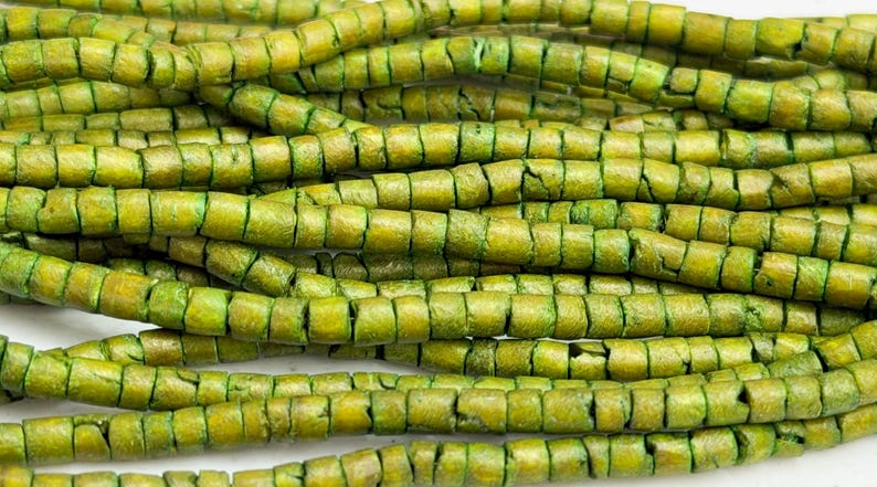 May include: Close-up of several strands of green beads. The beads are cylindrical with a square cross-section and have a textured, weathered appearance. The color is a mix of olive and chartreuse.