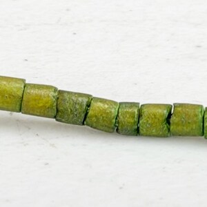 May include: A strand of olive green, square-shaped beads. The beads are strung together, creating a flexible line. The beads have a slightly weathered appearance, suggesting a vintage or handmade quality. The background is a plain white surface.
