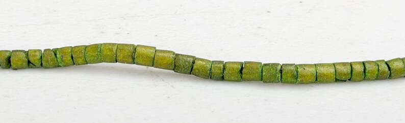 May include: A strand of olive green, square-shaped beads. The beads are strung together, creating a flexible line. The beads have a slightly weathered appearance, suggesting a vintage or handmade quality. The background is a plain white surface.