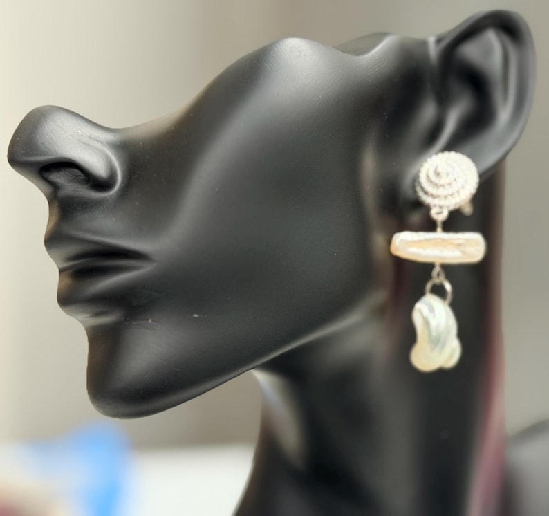 May include: A pair of earrings displayed on a black mannequin head. The earrings feature a silver spiral stud, a rectangular pearl, and a teardrop-shaped pearl pendant. The pearls have a creamy white color.