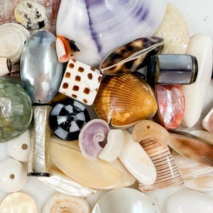 May include: A collection of assorted beads and pendants made from natural seashells and mother-of-pearl. The assortment includes various shapes, sizes, and textures, featuring iridescent white, brown, black, grey, and purple tones with patterns.