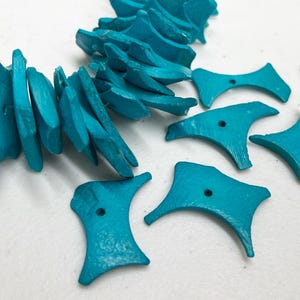 Puede incluir: Cuentas y elementos decorativos de madera de color turquesa. Las cuentas tienen forma irregular y están ensartadas, mientras que los otros elementos tienen un diseño geométrico único con un agujero en el centro. Los artículos están sobre una superficie blanca.