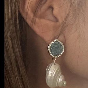May include: Earrings featuring a round, light blue stone surrounded by small, clear crystals. A white, shell-shaped pendant hangs below. The earrings are gold-toned and are shown against a blurred background.