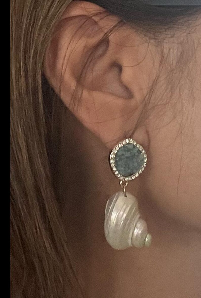 May include: Earrings featuring a round, light blue stone surrounded by small, clear crystals. A white, shell-shaped pendant hangs below. The earrings are gold-toned and are shown against a blurred background.