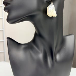 May include: A pair of seashell earrings displayed on a black mannequin. The earrings are pearlescent white and cream-colored, with a curved, spiral design. The earrings hang from silver-colored hooks.