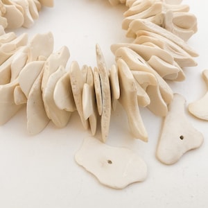 Puede incluir: Collar llamativo de color crema con cuentas de forma irregular y orgánica. El collar está compuesto por múltiples piezas, con algunas cuentas sueltas cerca. Las cuentas tienen un acabado mate y una estética natural y terrosa.